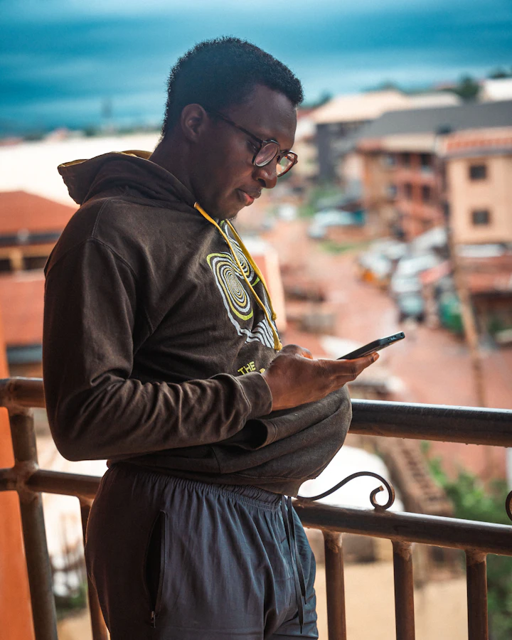 A man on a balcony checking his phone, with rooftops behind him